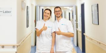 Das Bild zeigt eine Pflegekraft und einen Arzt, lächelnd mit einer Daumen-hoch-Bewegung