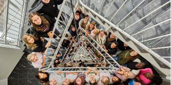 Neue Azubis August 2024 Jung Menschen stehen auf einer Treppe und schauen über mehrere Ebenen hinauf in die Kamera.