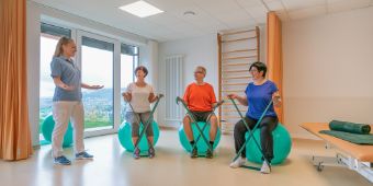 Eine Therapeutin führt Übungen mit Patienten durch, die auf Gymnastikbällen sitzen und Terrabänder halten