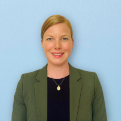 Text: Porträt von Daniela Krüger auf blauem Hintergrund, sie trägt einen grünen Blazer und ein schwarzes Shirt, ihre Haare sind zu einem Zopf gebunden und rotblond