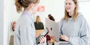 Das Foto zeigt zwei junge Frauen, die mit Macbook und Büchern in der Hand miteinander reden. 