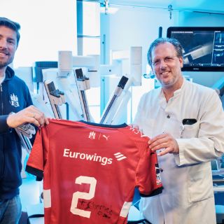 Prof. Dr. Christian Wülfing (rechts) und Tobias Schimmelbauer (links) präsentieren das Trikot, das der Handballer mit dem Operationsroboter signiert hat.