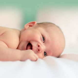 fEin Baby liegt auf dem Bauch und lächelt in die Kamera. Der Fokus liegt auf dem Kind. Der Hintergrund ist verschwommen.