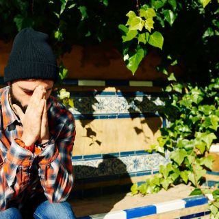 Auf einer Bank im grünen Sicht eine Person, welche die Hände vor dem Gesicht zusammengeschlagen hat