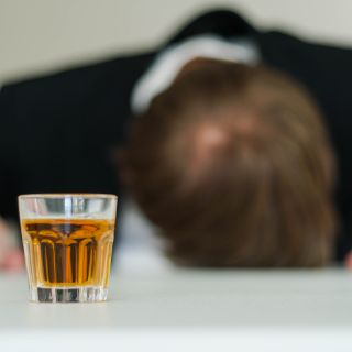 Im Vordergrund ist ein gefülltes Glas zu sehen. Im Hintergrund liegt eine Person mit dem Kopf auf dem Tisch. 