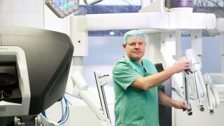 Das Bild zeigt Prof. Daniel Perez in OP-Kleidung. Er hält einen Intrumentenarm des Operationsroboters im leeren OP-Saal.