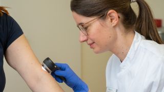 Das Motiv zeigt eine Dermatologin beim Untersuchen eines Armes.