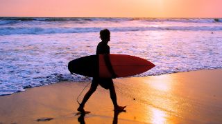 Das Bild zeigt einen Surfer mit Surfbrett unter dem Arm am Strand im Sonnenuntergang.