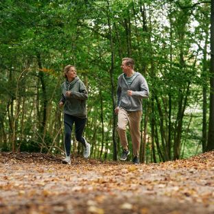 Zwei Menschen joggen lächelnd nebeneinander durch einen Wald