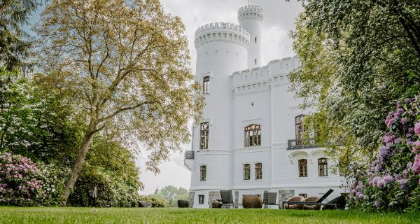 Ansicht der Blomenburg aus dem Gartenareal Eine weiße Burg umgeben von prächtigen Grün und rosa Blumen