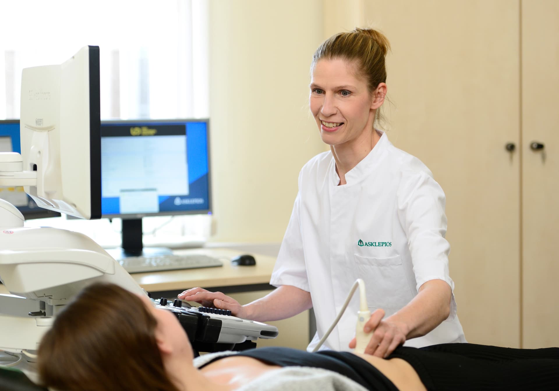 Text: Dr. Simone Klübe bei Ultraschalluntersuchung mit einer Patientin. Sie hat blonde Haare zu einem Dutt gebunden und trägt einen weißen Kittel