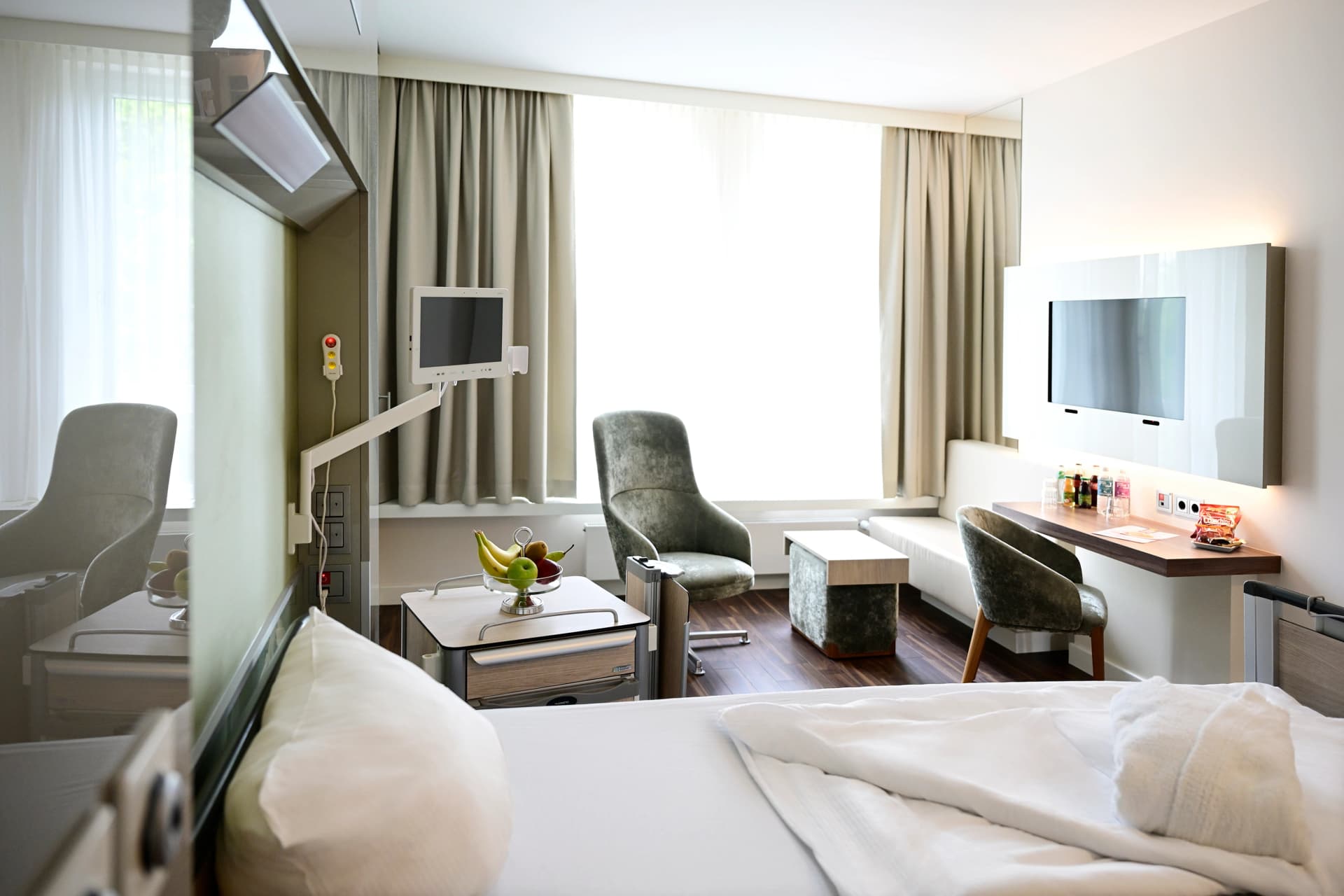 Helles, modern eingerichtetes Patientenzimmer mit großem Fenster, Bett, gemütlichem Sessel, kleinem Tisch und Schreibtischbereich.