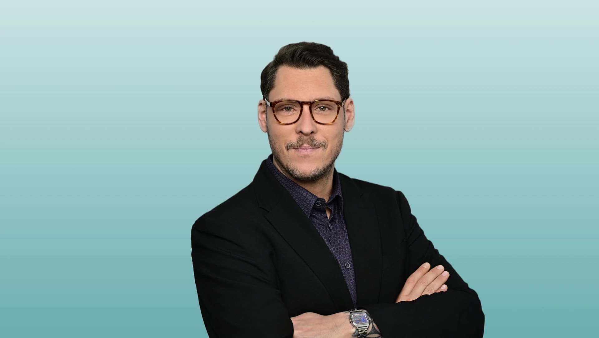 Bild: Portrait eines Mannes mit Brille und verschränkten Armen vor hellblauem Hintergrund. Er trägt ein dunkles Sakko und blickt direkt in die Kamera.