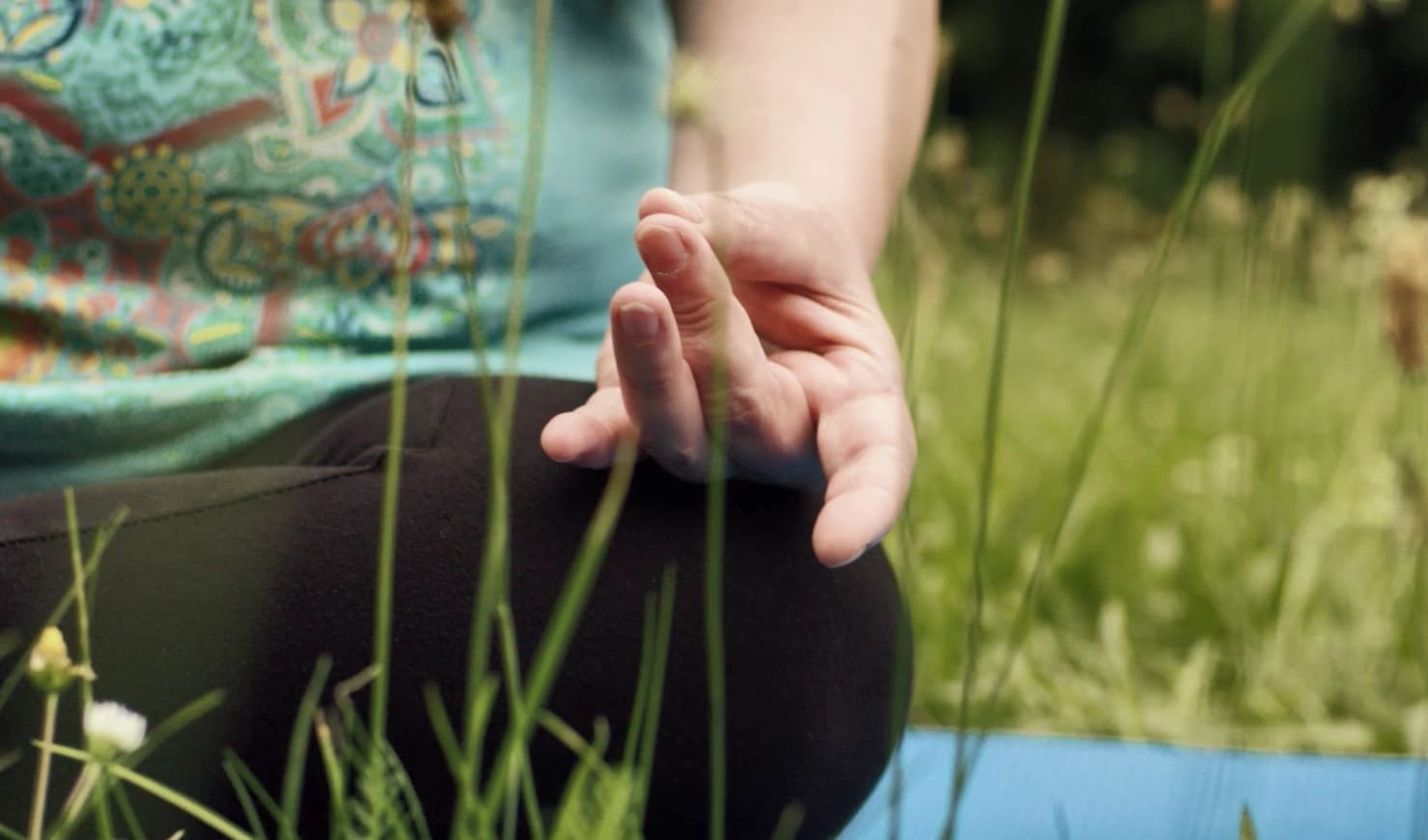 Das Bild zeigt eine Person im Gras im Schneidersitz sitzend und die Finger in Meditationshaltung