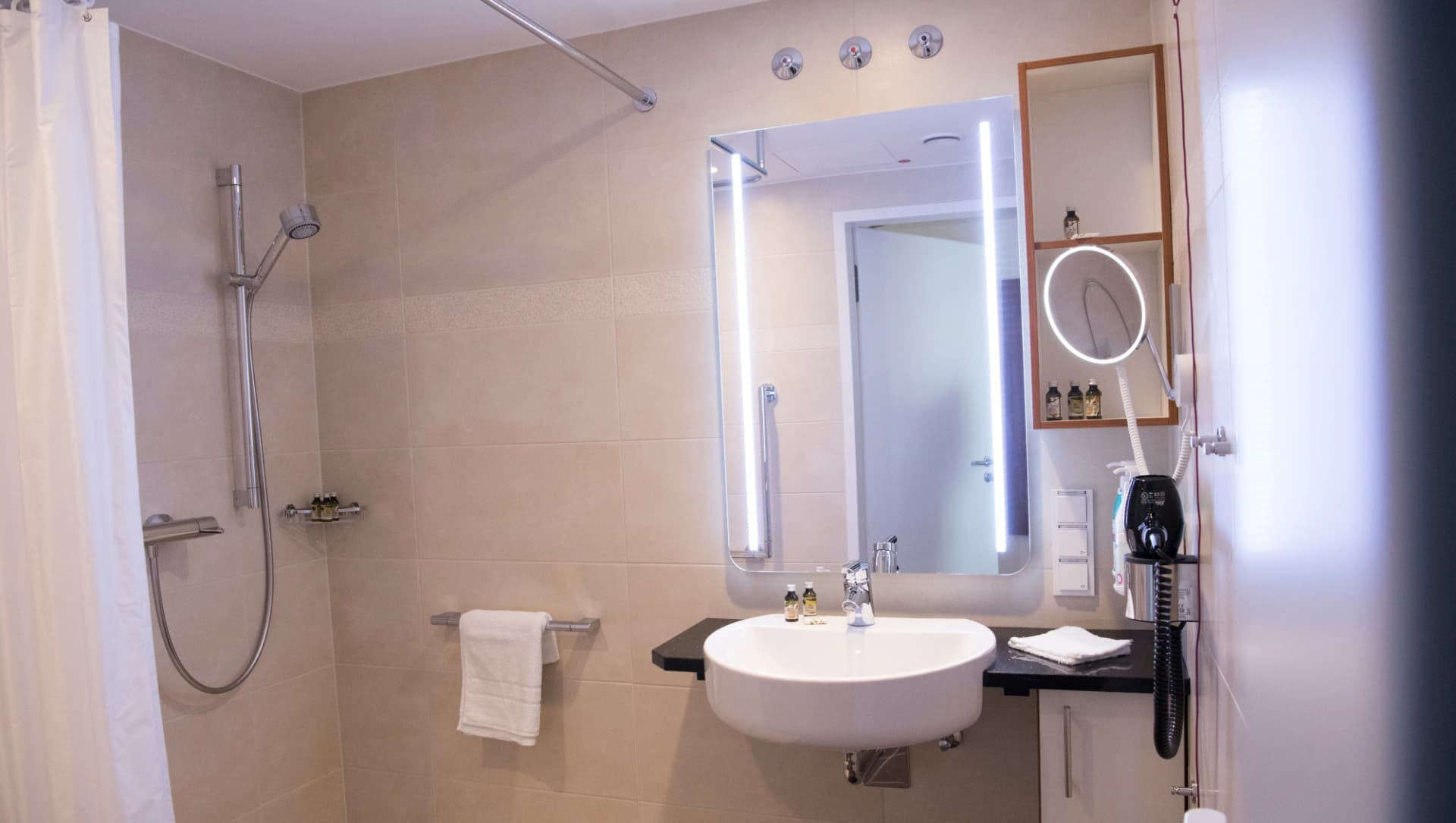 Das Bild zeigt ein hellbeiges Badezimmer auf der Privatstation mit Dusche, Waschbecken, beleuchtetem Spiegel, Schmickspiegel und Handtüchern