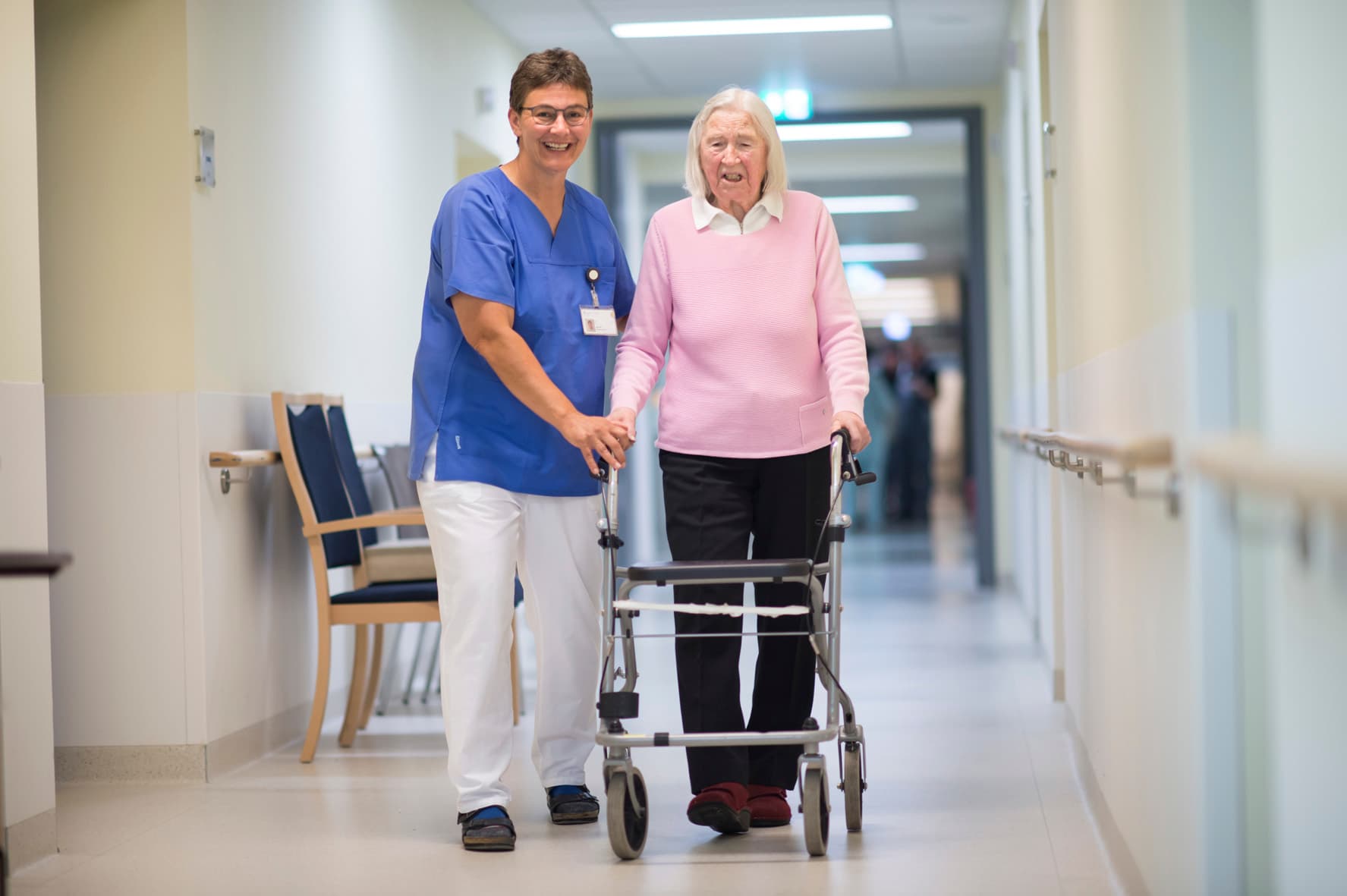 Das Motiv zeigt eine Pflegekraft mit einer älteren Dame am Rollator