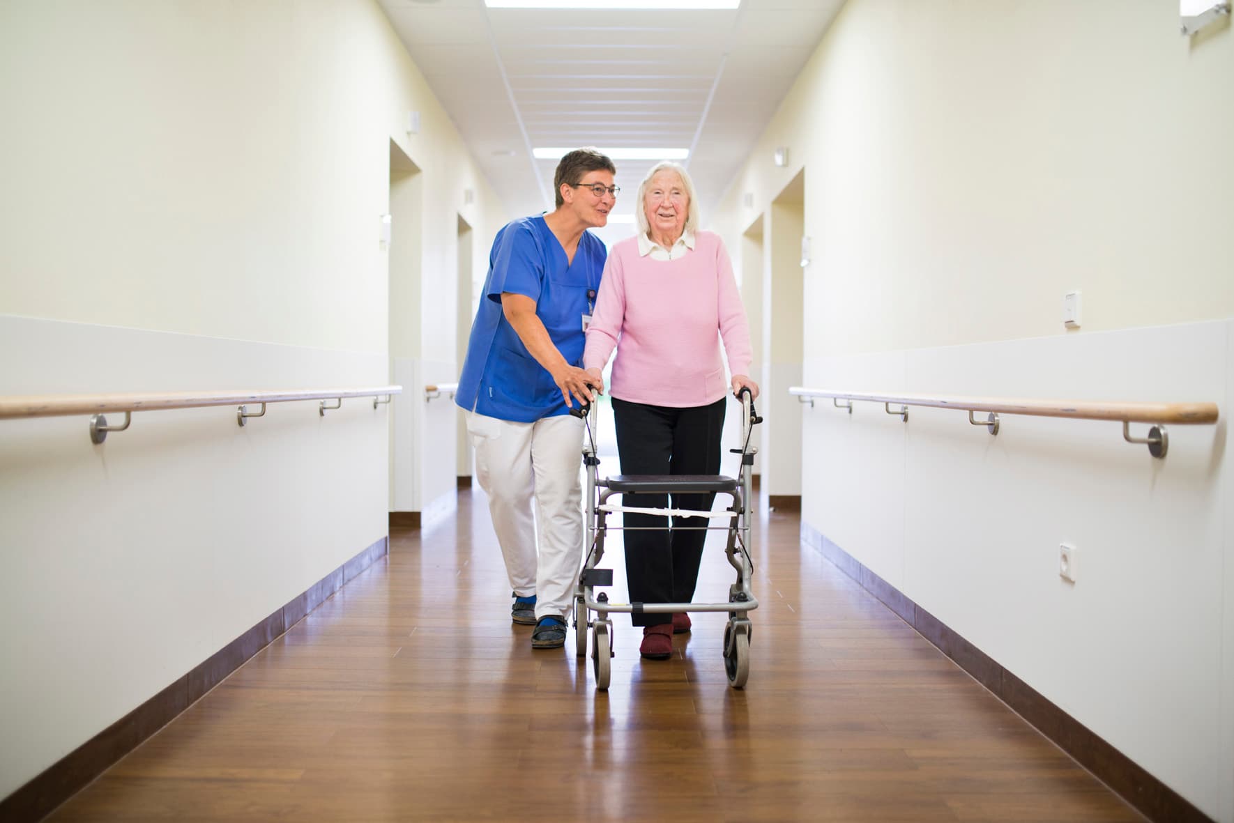 Das Motiv zeigt eine alte Dame am Rollator mit einer Pflegekraft