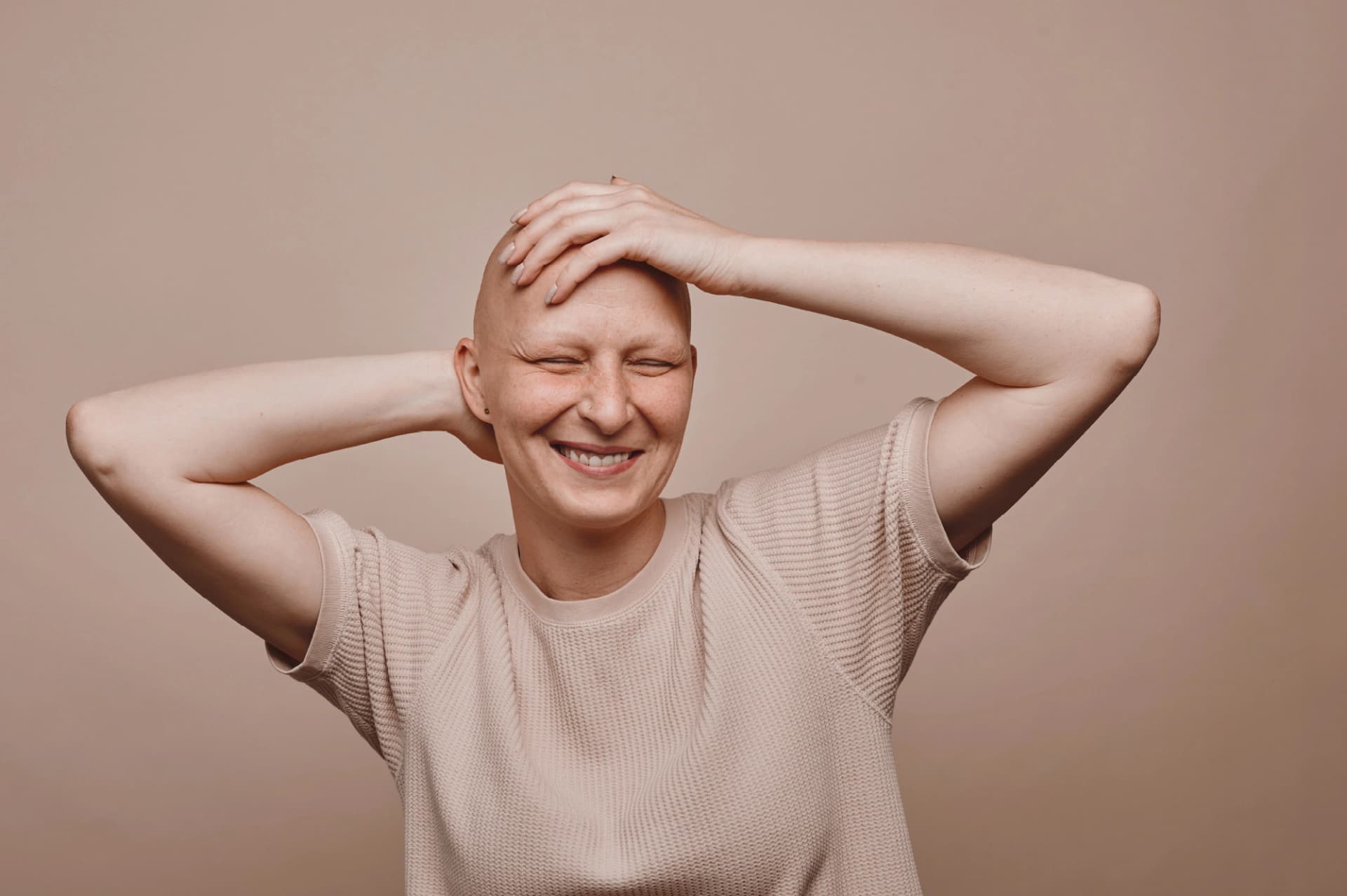 Portrait einer lächelnden Frau in Beige-Tönen ohne Haare, die sich glücklich mit der Hand auf den Kopf fasst.