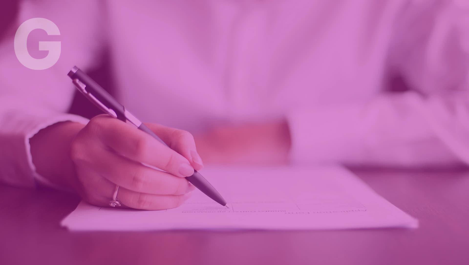 Hand mit einem Ring hält einen Stift und schreibt auf weißes Papier auf einem Tisch.