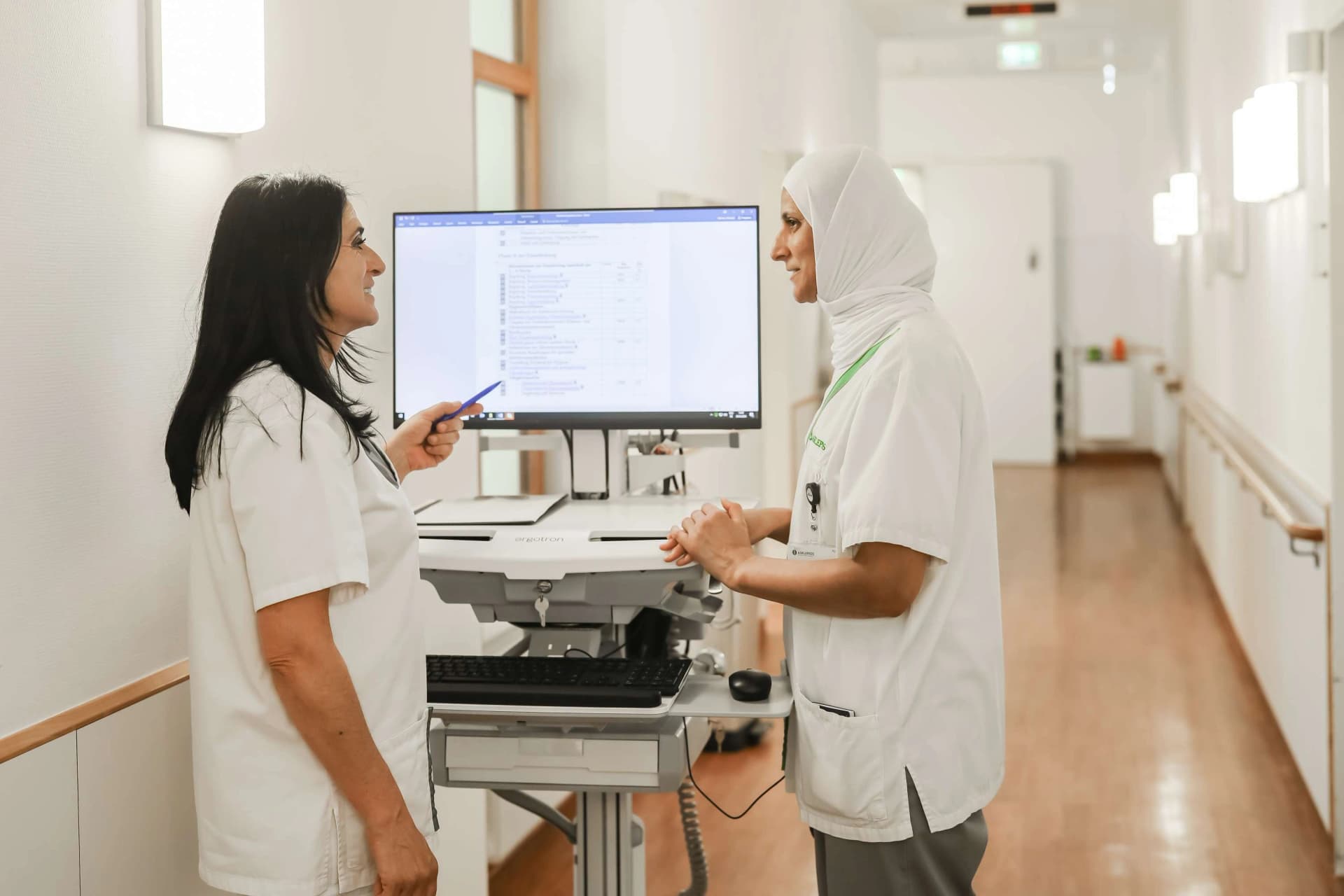 Das Motiv zeigt wie eine internationale Auszubildende von einer examinierten Fachkraft in das Dokumentationssystem eingewiesen wird.