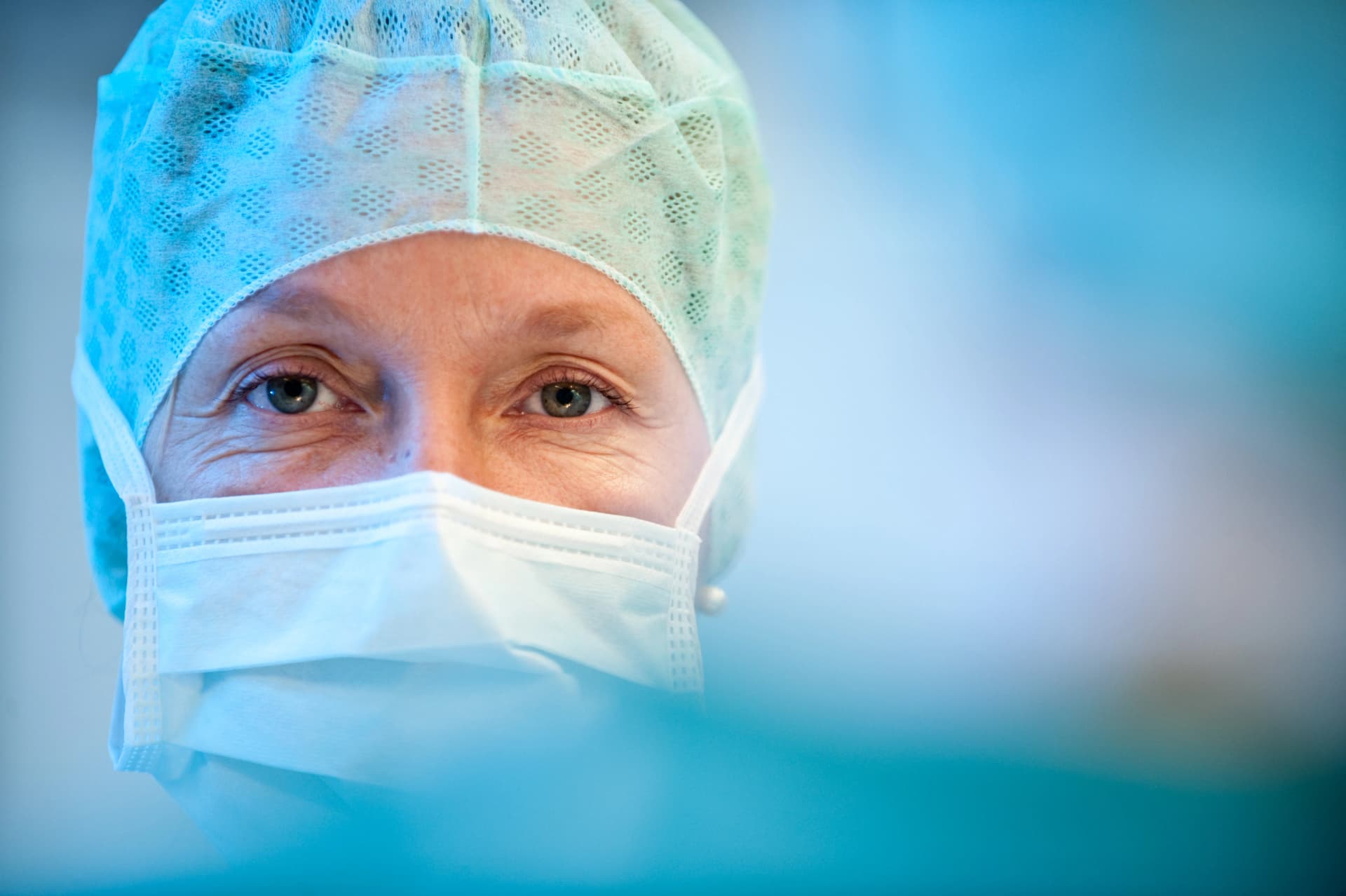 Das Bild zeigt eine Nahaufnahme einer weiblichen Chirurgin in OP-Kleidung. Ihre Augen sind freundlich. Sie trägt einen Mund-Nasen-Schutz und eine OP-Haube.