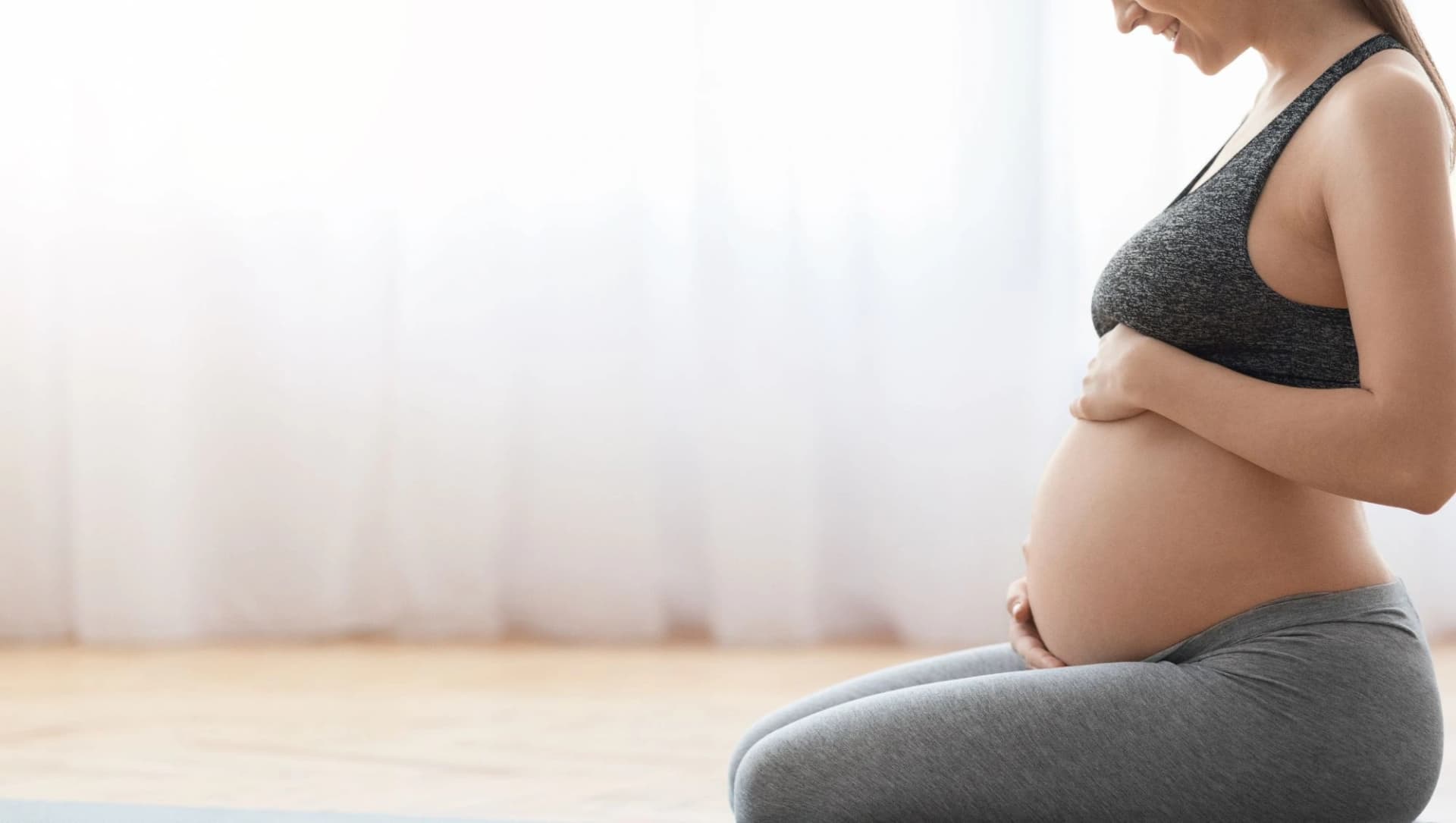 Eine schwangere Frau sitzt in Sportkleidung auf einer Turnmatte und hält liebevoll ihren Bauch – die Frau lächelt.