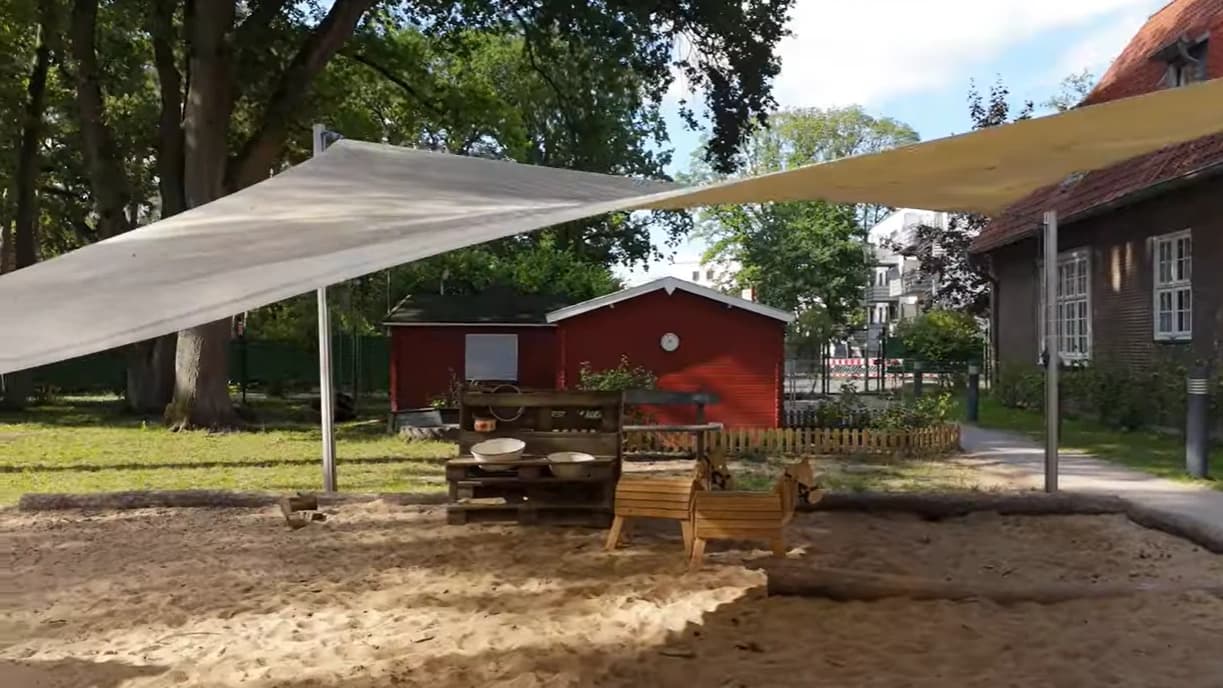 Bild: Spielküche im Sandkasten mit Sonnensegel und rotem Schuppen im Hintergrund