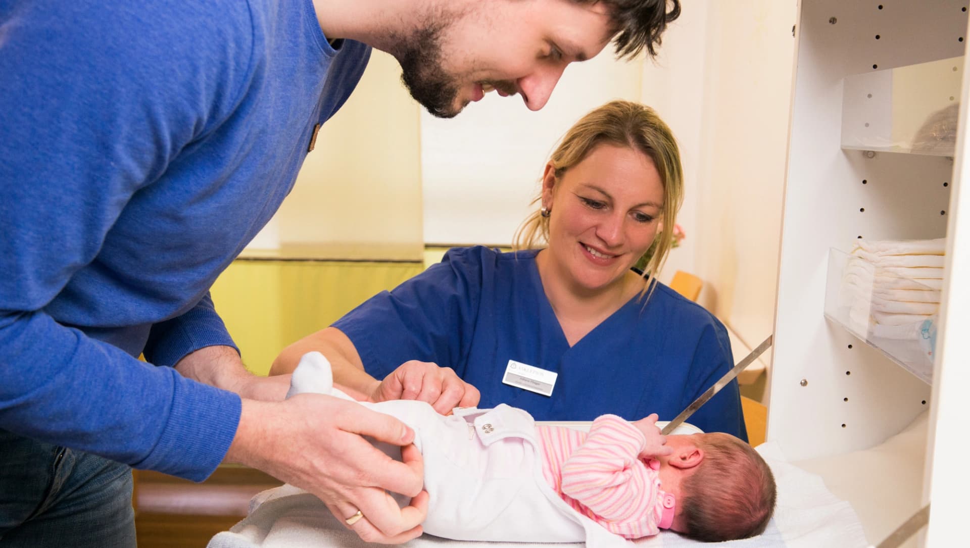 Das Bild zeigt einen Vater, der sein Neugeborenes unter Anleitung einer Pflegeexpertin versorgt