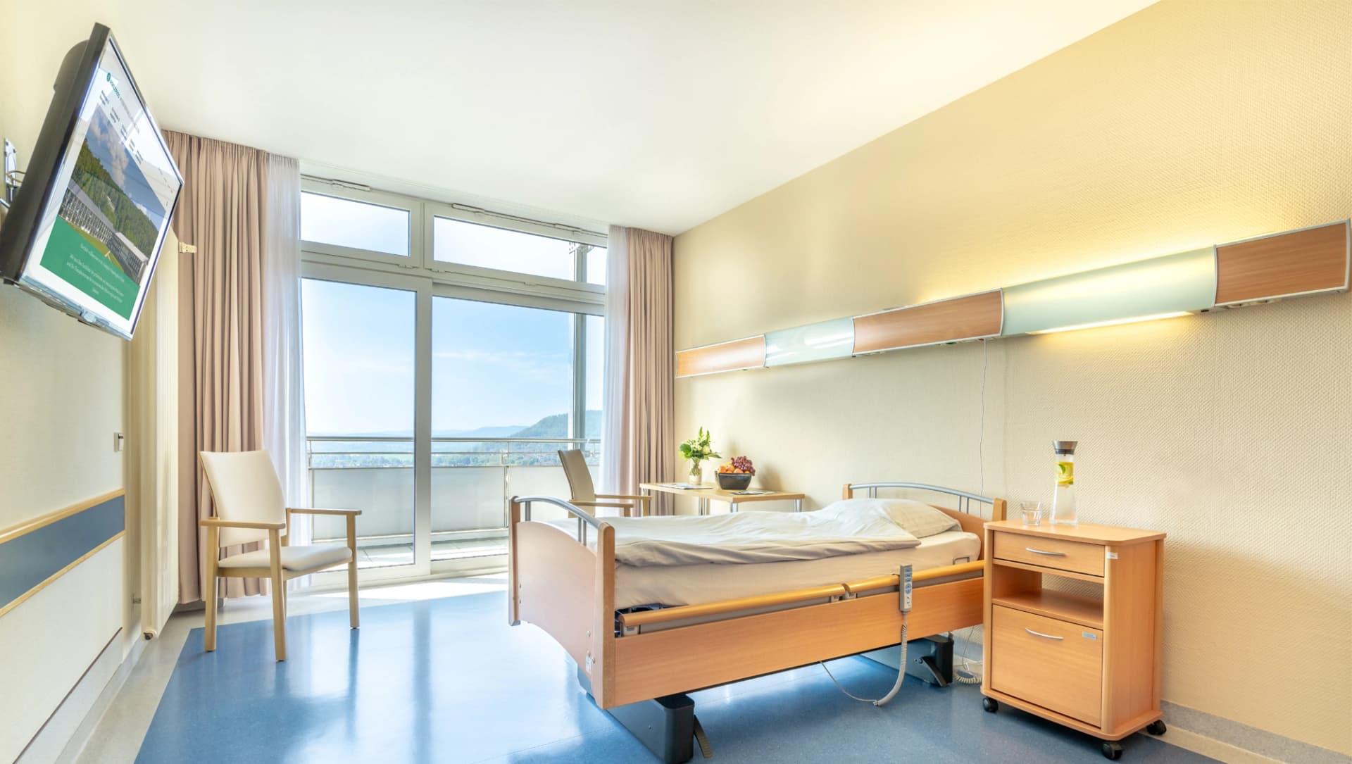 Eine Darstellung eines Patientenzimmers mit Bett, Nachtschrank, TV und Stuhl