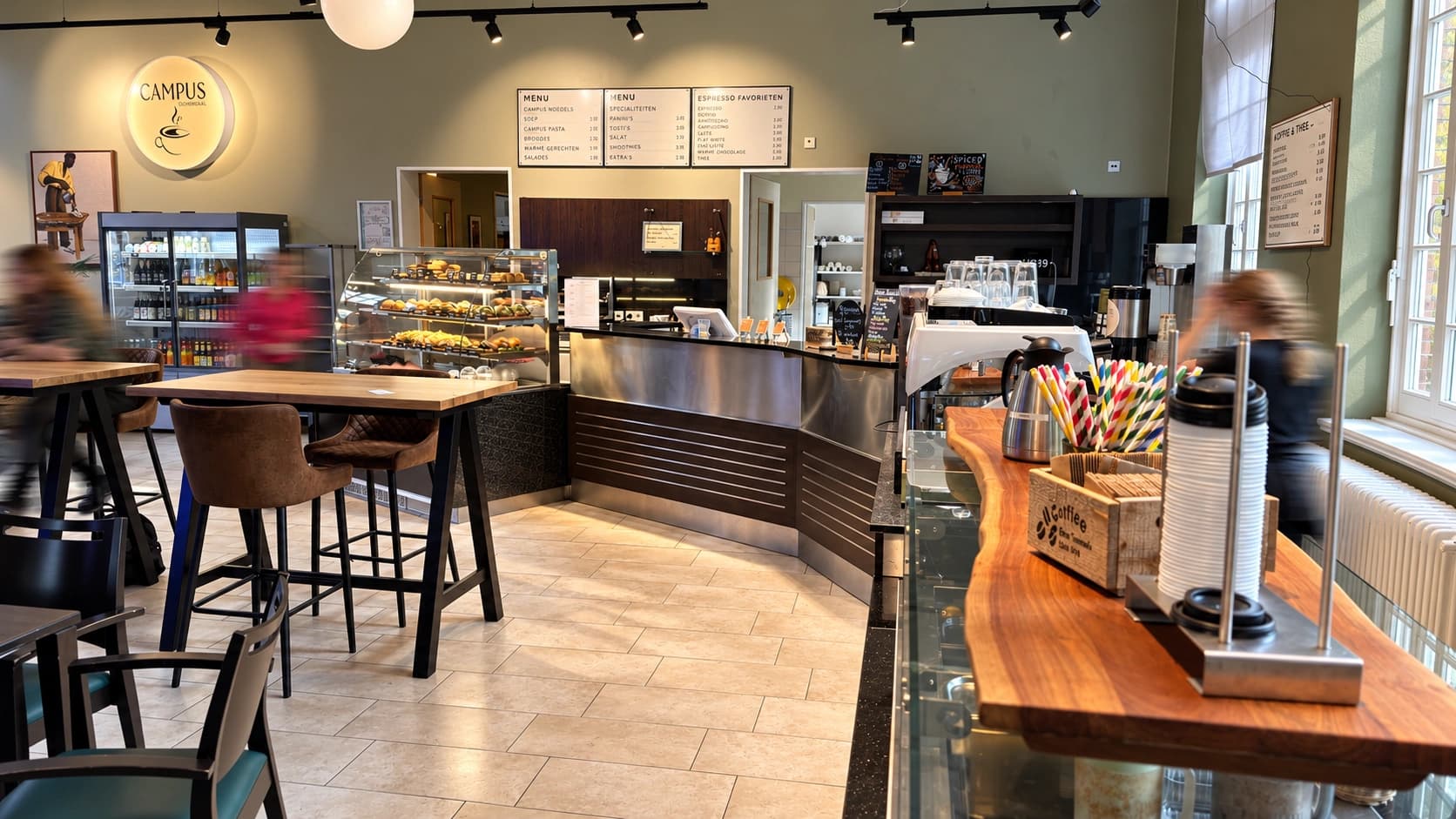 Innenbereich des Campus-Cafés Ochsenzoll mit Kaffeebar, Kuchenvitrine, Sitzgelegenheiten und bewegungsunscharfen Personen.