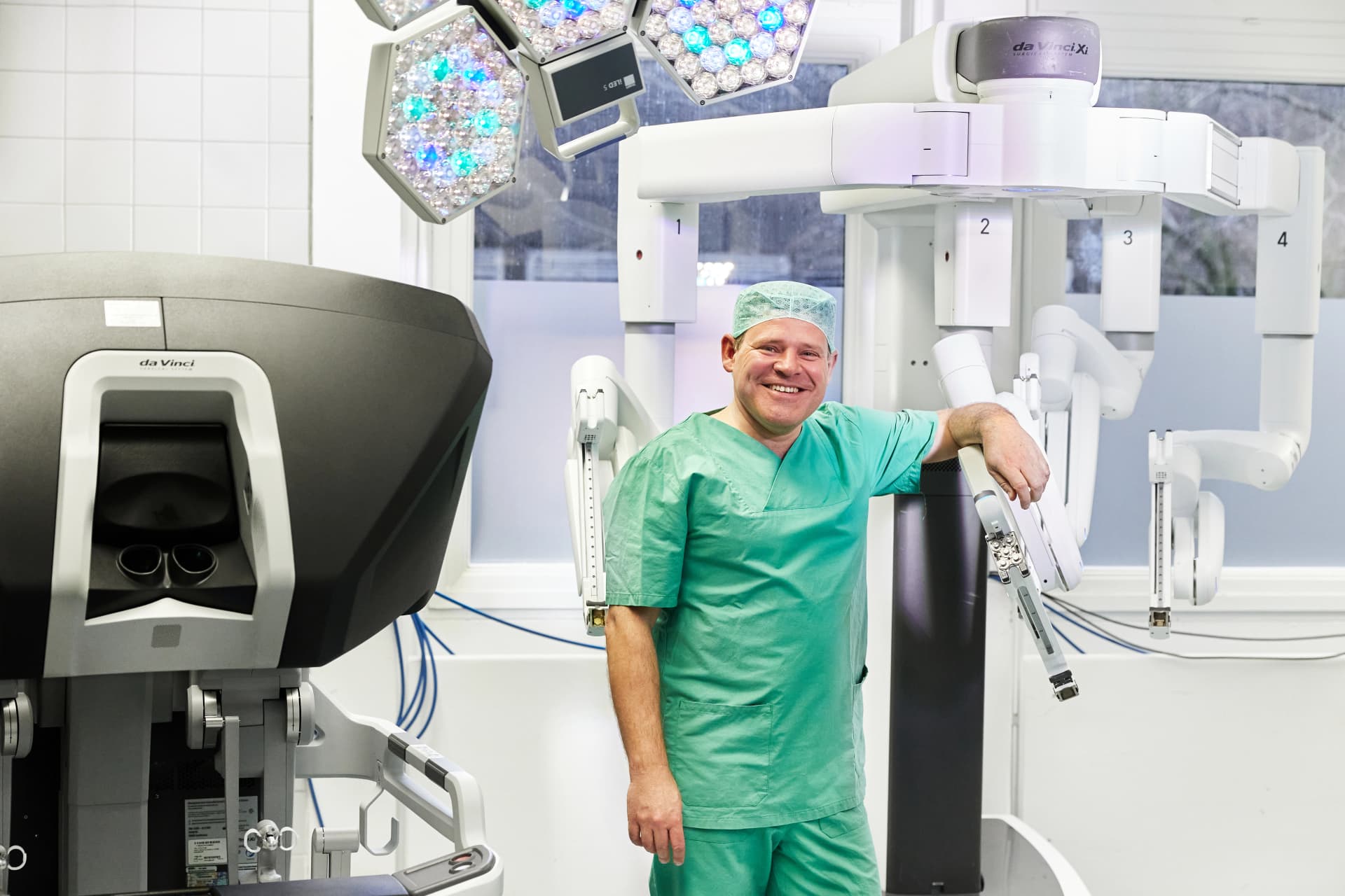 Das Bild zeigt Prof. Daniel Perez in OP-Kleidung. Er hält einen Intrumentenarm des Operationsroboters im leeren OP-Saal.