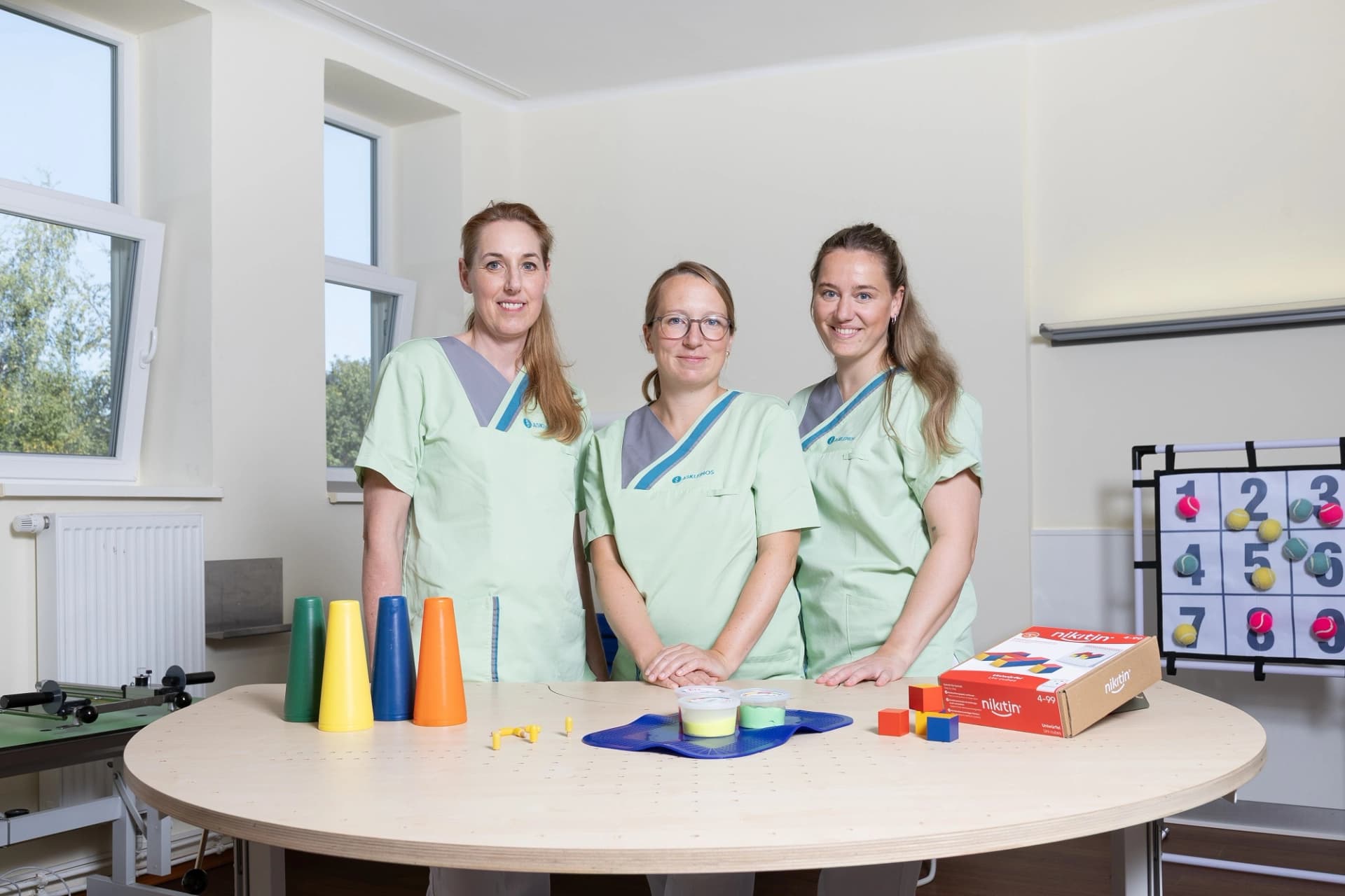 Das Bild zeigt die drei Ergotherapeutinnen vor einem halbrunden Arbeitstisch mit verschiedenen Materialien, die in der Therapie verwendet werden können