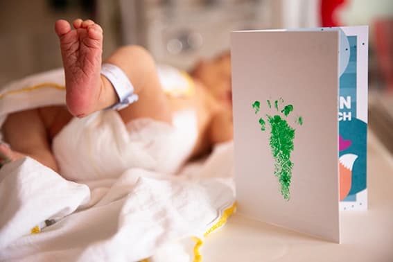 Das Motiv zeigt ein Neugeborenes und daneben steht eine Glückwunschkarte mit einem Babyfußabdruck.