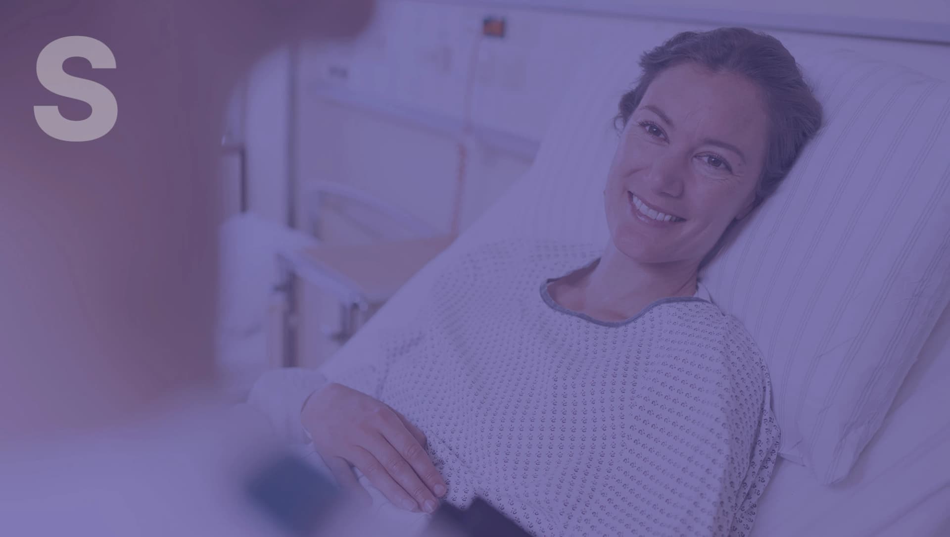 Lächelnde Person in einem Krankenhausbett trägt weißes Krankenhaushemd mit Muster, eine unscharfe Person im Vordergrund.