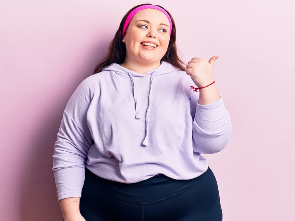 Das Motiv zeigt eine junge, lächelnde Frau in einem violetten Kaupuzenpullover, die vor einer rosa Wand steht und mit dem Daumen in eine Richtung zeigt.