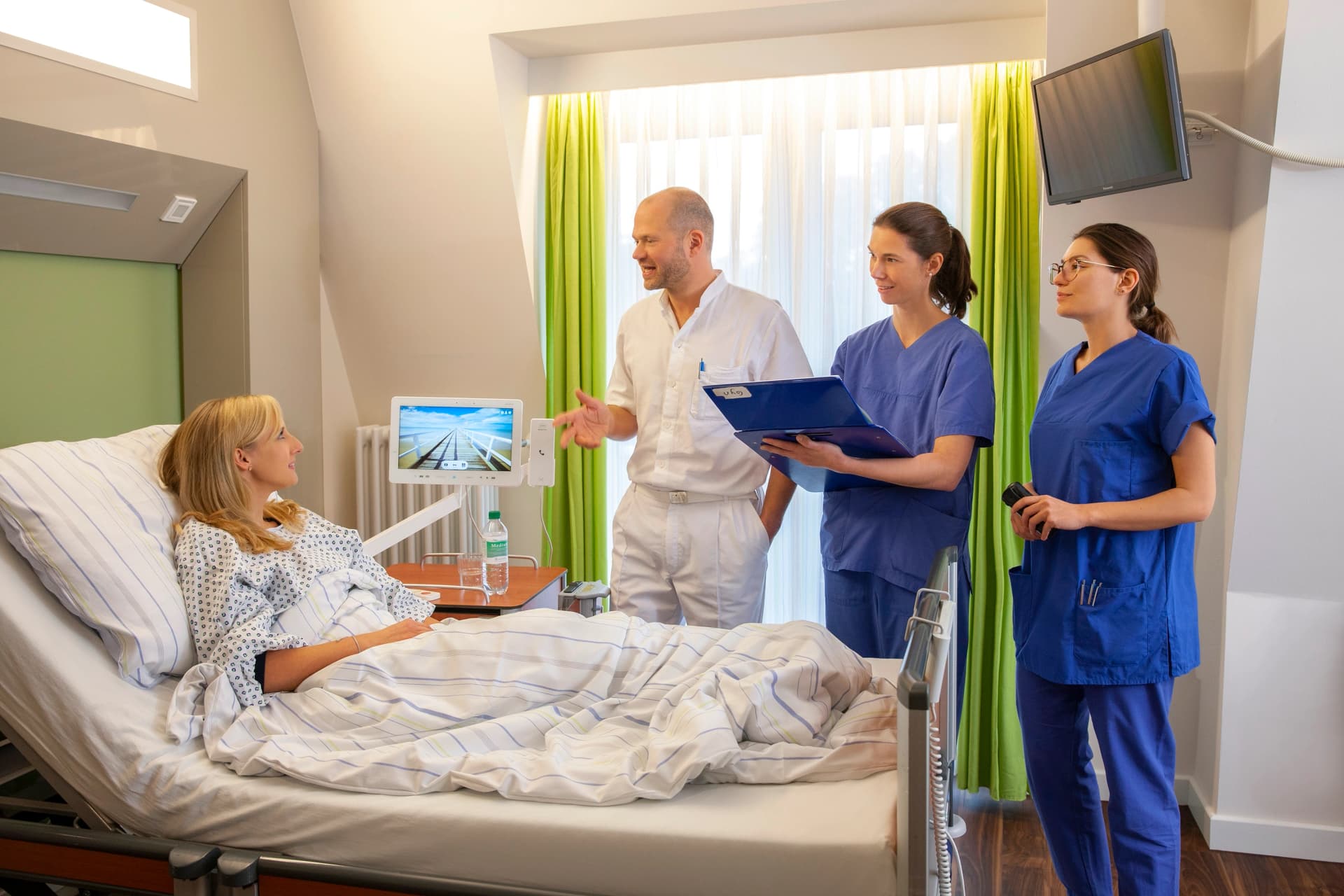Das Motiv zeigt Chefarzt Dr. med. Wolf und zwei Assistentinnen an einem Patienbett zur Visite.
