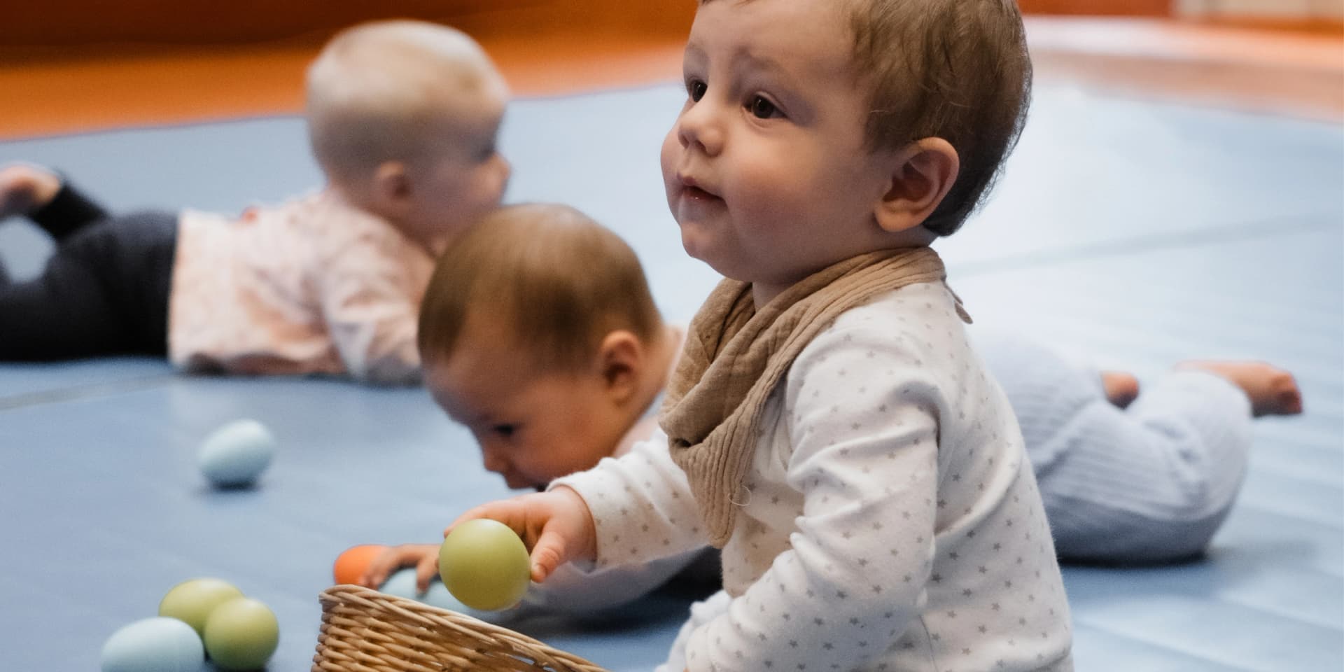 Nahaufnahme eines Kindes das im Kurs zu seiner Mama schaut. Es hält in der Hand ein Rasselei. Im Hintergrund krabbeln zwei andere Kinder über eine blaue Turnmatte.