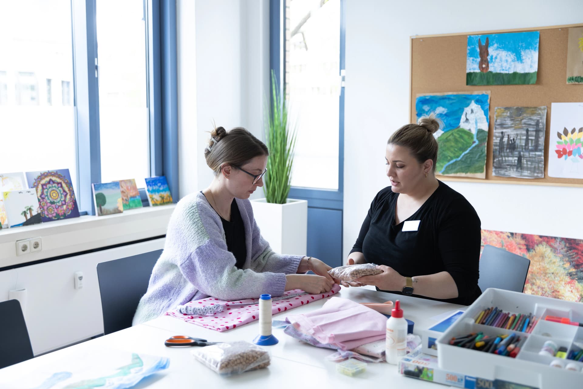 Das Motiv zeigt zwei Mitarbeiterinnen der Schmerz-Tages-Klinik, die im Kreativraum an einem Tisch sitzen und die Therapie vorbereiten.