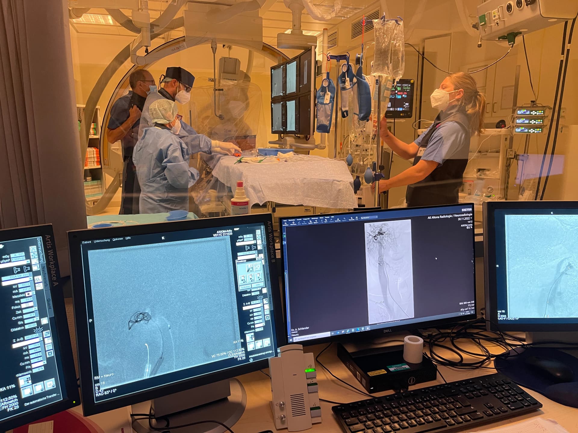 Blick aus dem Kontrollraum in die Angiografie. Dort wird gerade von Kolleg:innen eine Intervention vorbereitet.