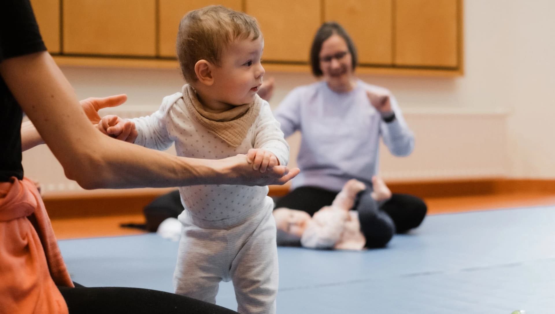 Ein Baby hält sich bei einem Kurs an den Händen seiner Mama fest. Im Hintergrund sieht man eine singende Mama mit ihrem liegenden Kind.