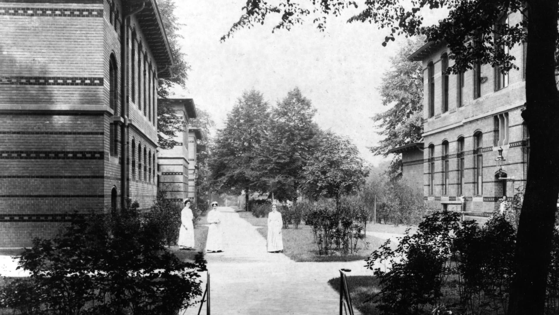 Das Foto zeigt Frauen, die in einem Garten zwischen Häuserfronten des Allgemeinen Krankenhauses stehen. Die Aufnahme wurde um 1906 gemacht.